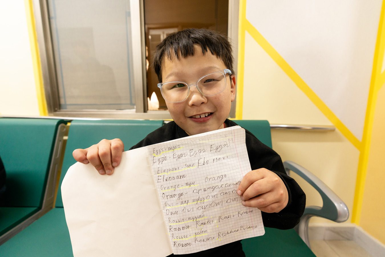 A boy with glasses smiles and shows his notebook of English words