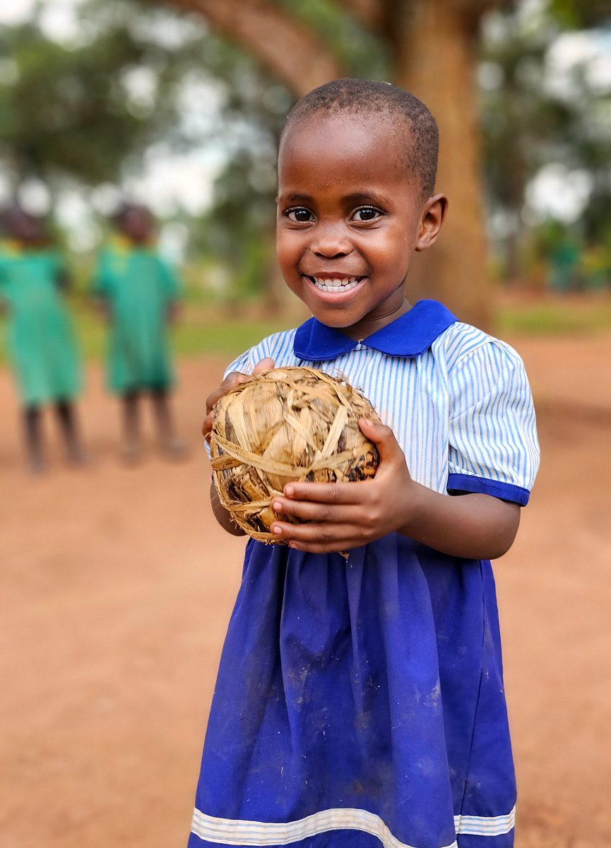 Little girl in blue and white school uniform holds ball at Wakivule school on National Play Day in Uganda