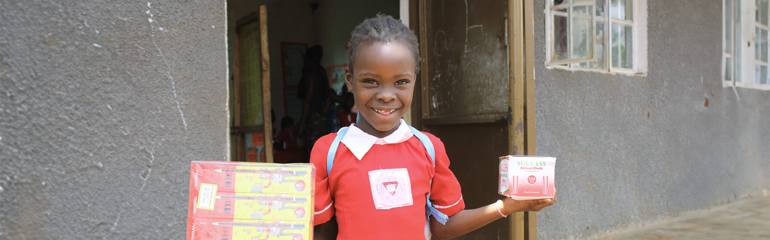 School age girl standing outside her classroom, smiling and holding supplies