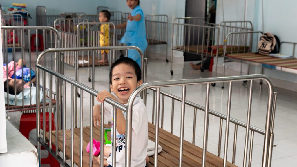 A boy growing up in an orphanage in Vietnam sits in a metal slat crib without any blankets or comfort items.