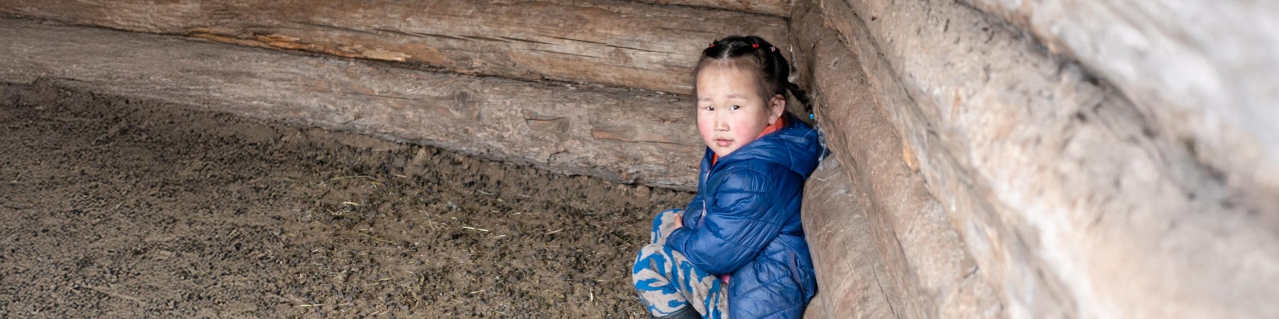 Little girl in Mongolia wearing a blue jacket sitting outside, leaning up against the barn wall