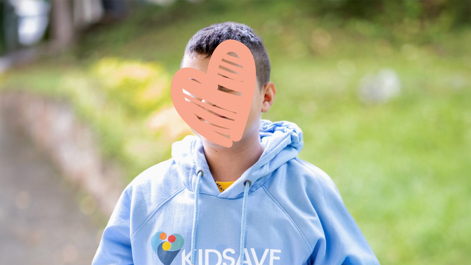 A waiting child wears a blue sweatshirt outdoors. His face is covered by a heart for privacy.